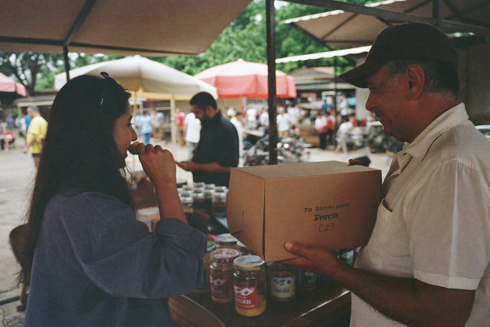 Produtor entregando uma caixa de bolo para uma cliente numa barraca de feira, com potes de doce na mesa.